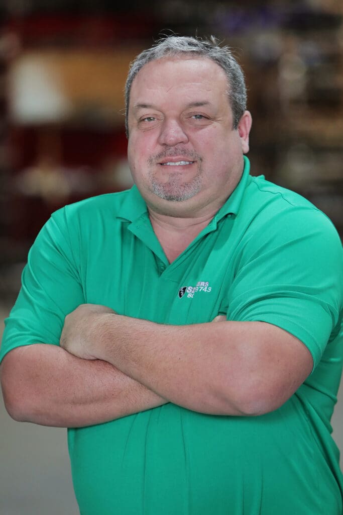 A man with a light complexion and short, graying hair stands with his arms crossed, smiling slightly. He is wearing a bright green polo shirt