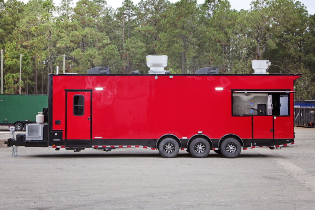 A red food or BBQ trailer with multiple axles and exhaust vents on the roof, parked outdoors with trees in the background.
